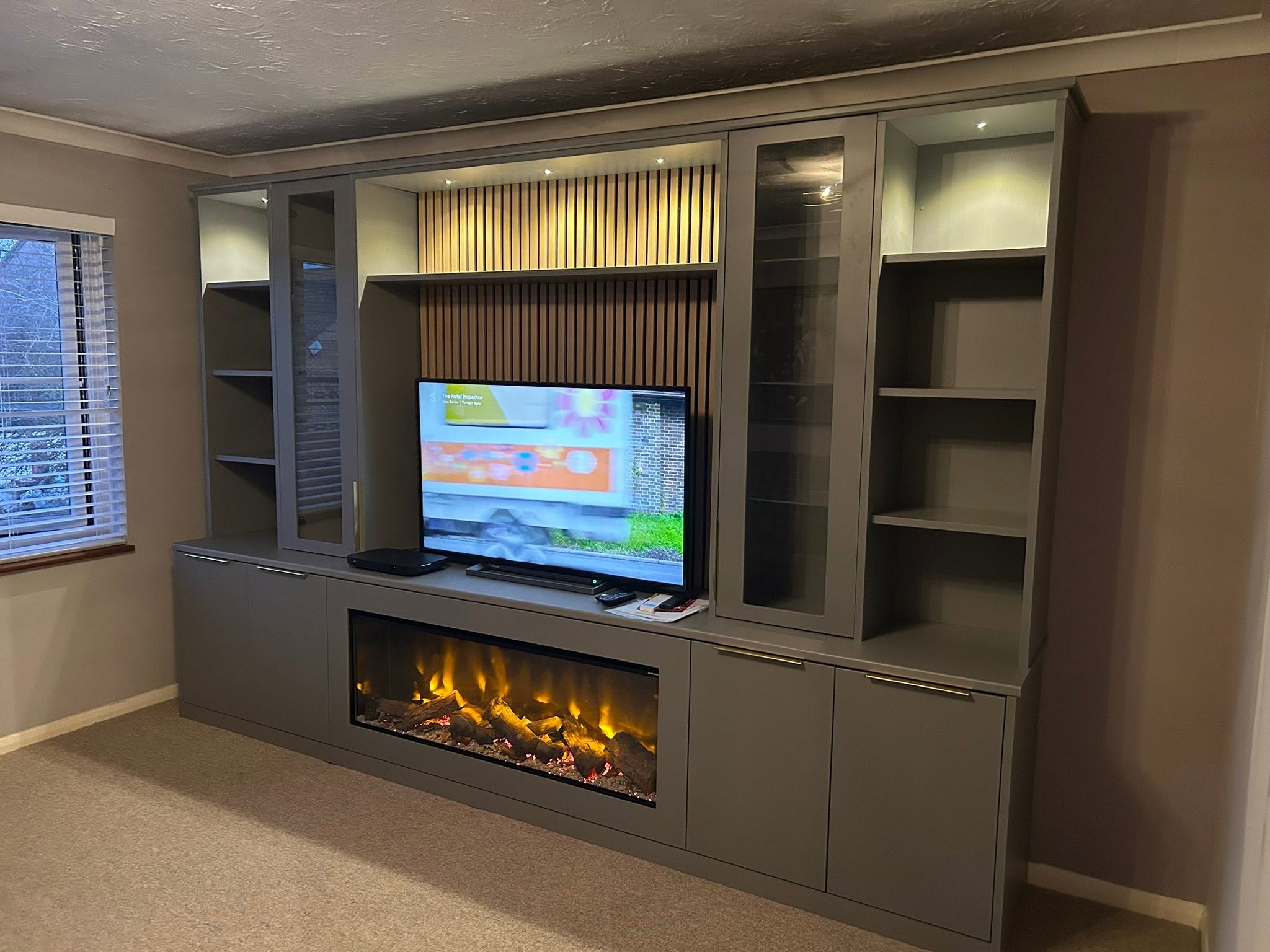 Grey fitted media wall with TV, electric fire, glass cabinets and open shelving